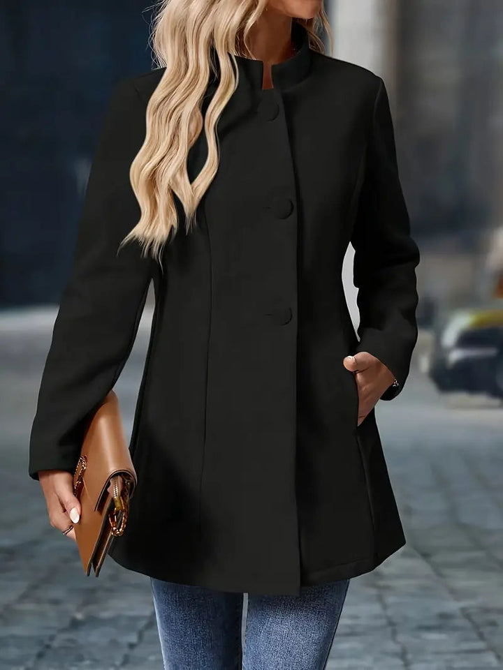 Black mid-length coat with a stand collar, tailored waist, and covered button front, worn with blue jeans.