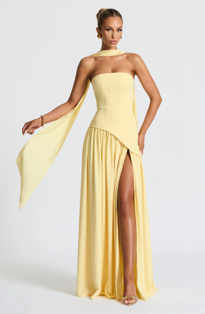 Model wearing a pale yellow strapless corset maxi dress with a high leg slit and elegant draped scarf detail.