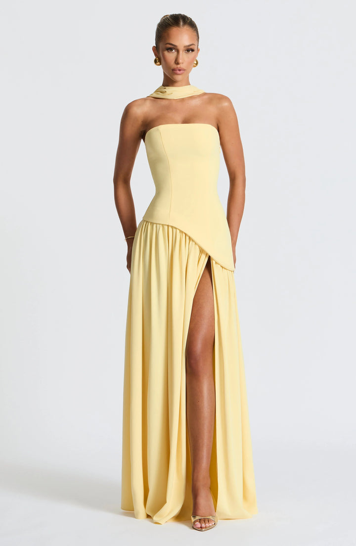 Model wearing a pale yellow strapless corset maxi dress with a high leg slit and draped scarf detail.