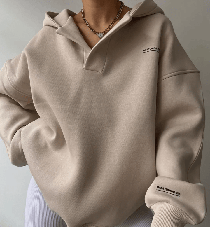 Beige oversized V-neck pullover hoodie with drop shoulders, ribbed cuffs, and small black text branding on chest and sleeve.