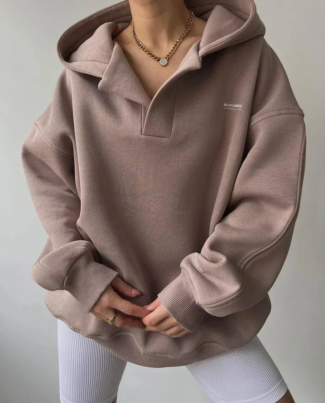 Taupe oversized V-neck hoodie with drop shoulders, ribbed cuffs, and subtle chest branding, worn over white leggings.