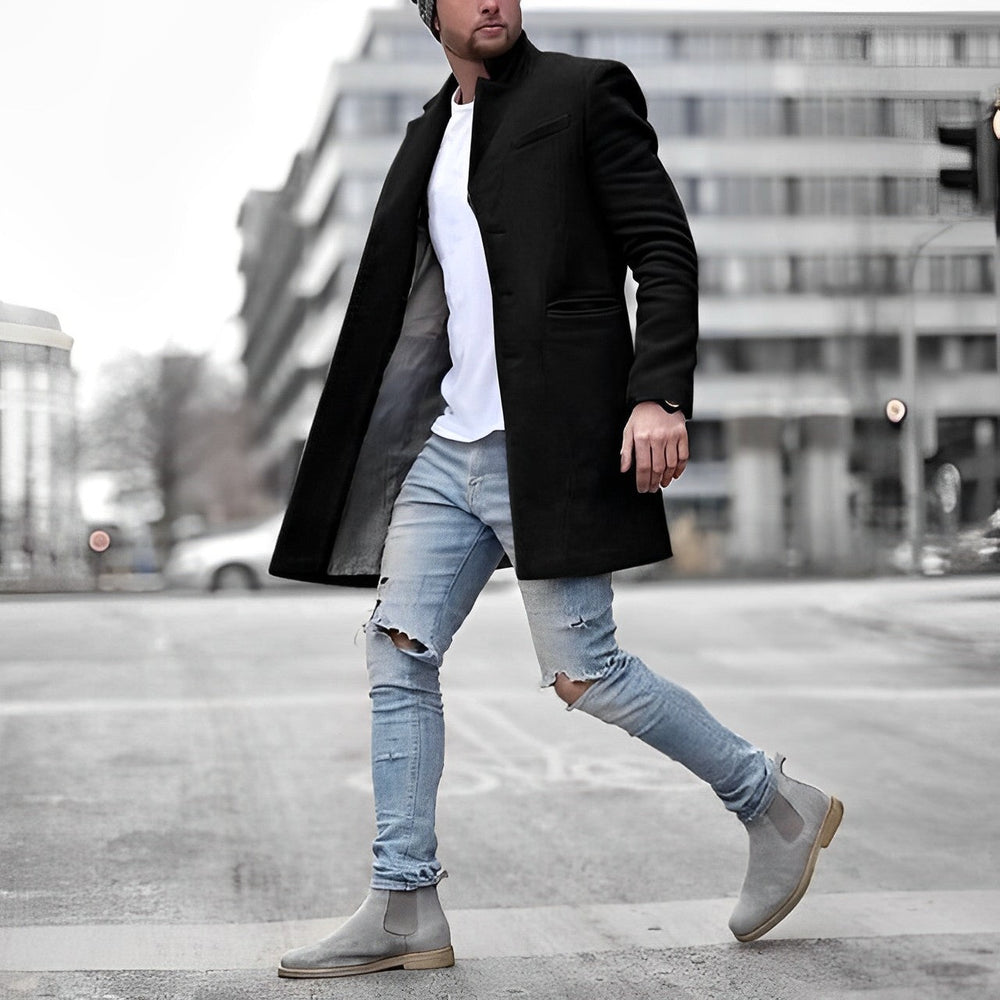 Man wearing a black slim-fit stand collar overcoat over a white t-shirt, light blue distressed jeans, and grey suede boots.