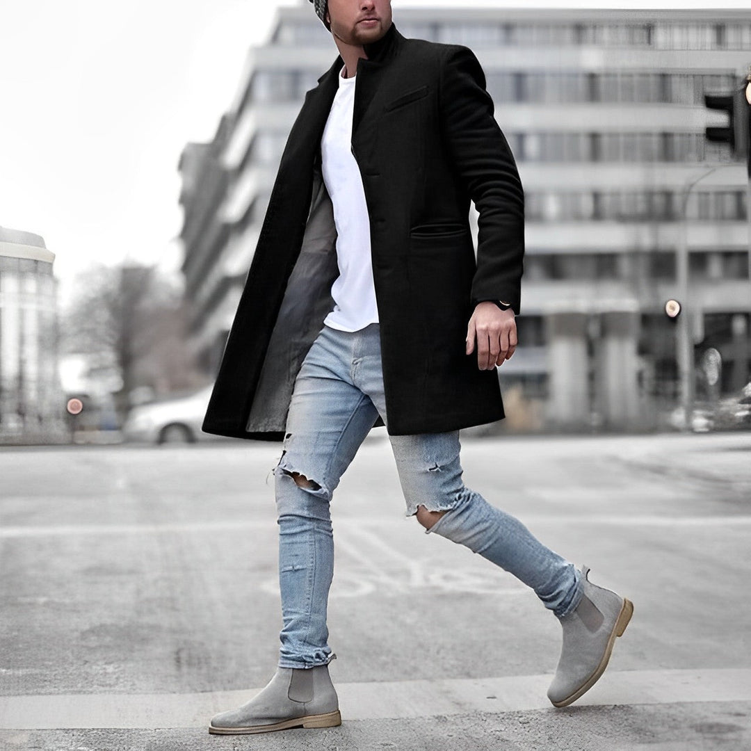 Man wearing a black slim-fit stand collar overcoat over a white t-shirt, light blue distressed jeans, and grey suede boots.