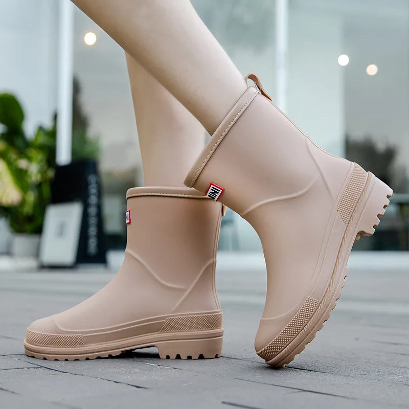 Beige mid-calf waterproof rain boots with a grippy treaded sole and back pull tab worn on a paved walkway.