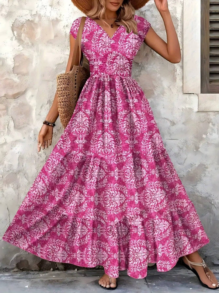 Pink floral boho maxi dress with a V-neckline, smocked bodice, and tiered skirt, styled with a straw tote and sandals.